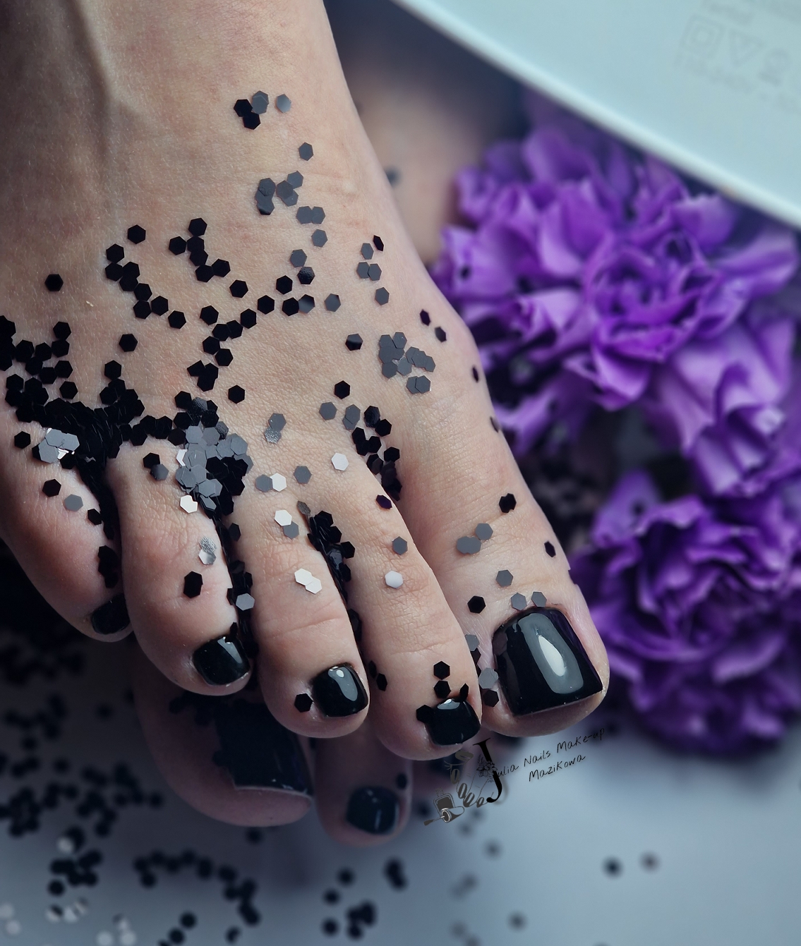 Zwarte pedicure met zilveren confetti naast paarse bloemen